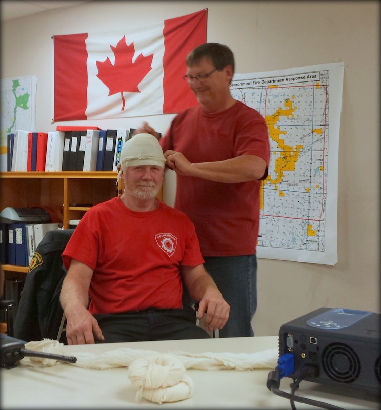 Volunteer practising a head bandage on a seated member at the fire hall, with the response-area map behind