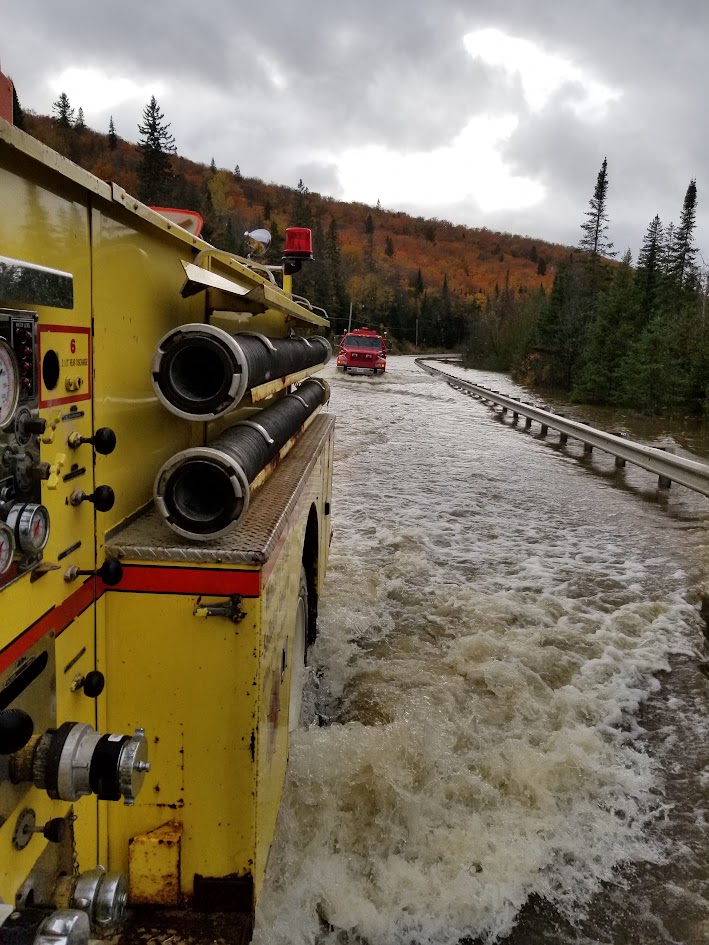 View from the side of a yellow pumper wading through fast floodwater behind another apparatus