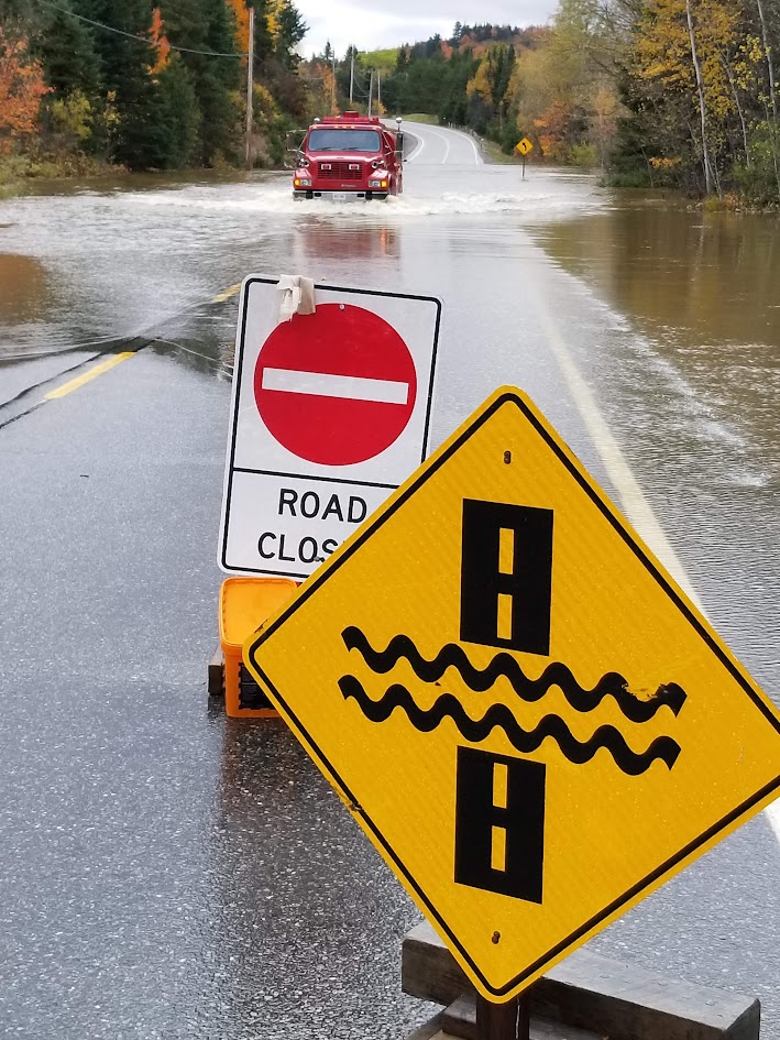 Road Closed and flooded-road signs with a pumper driving through floodwater behind them