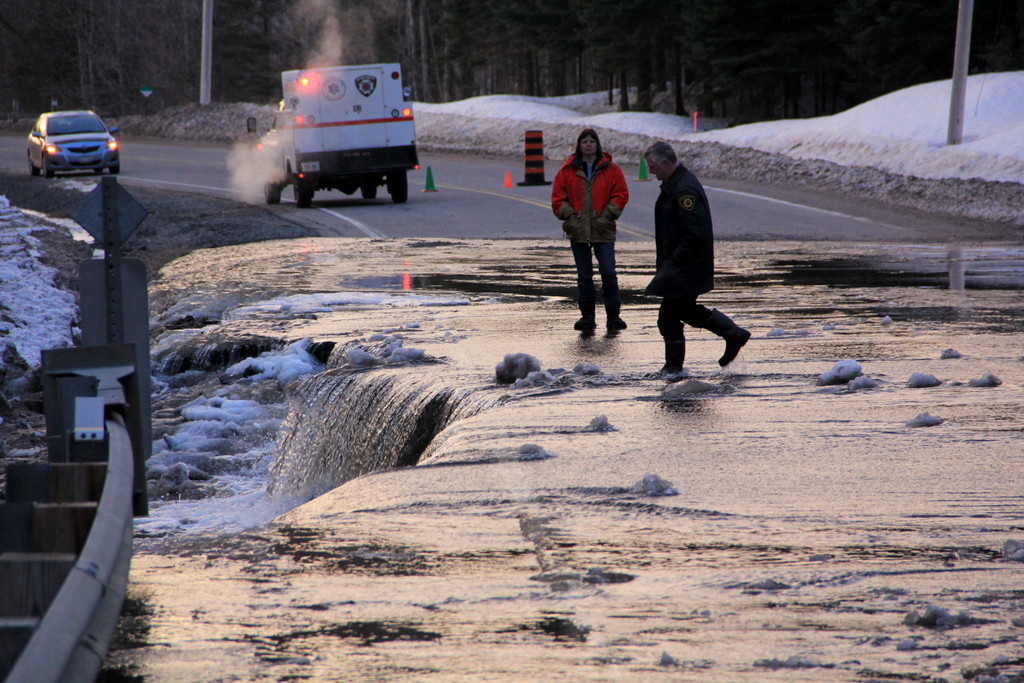 Meltwater pouring over a road edge at dusk while responders check the flooded section and an emergency vehicle waits