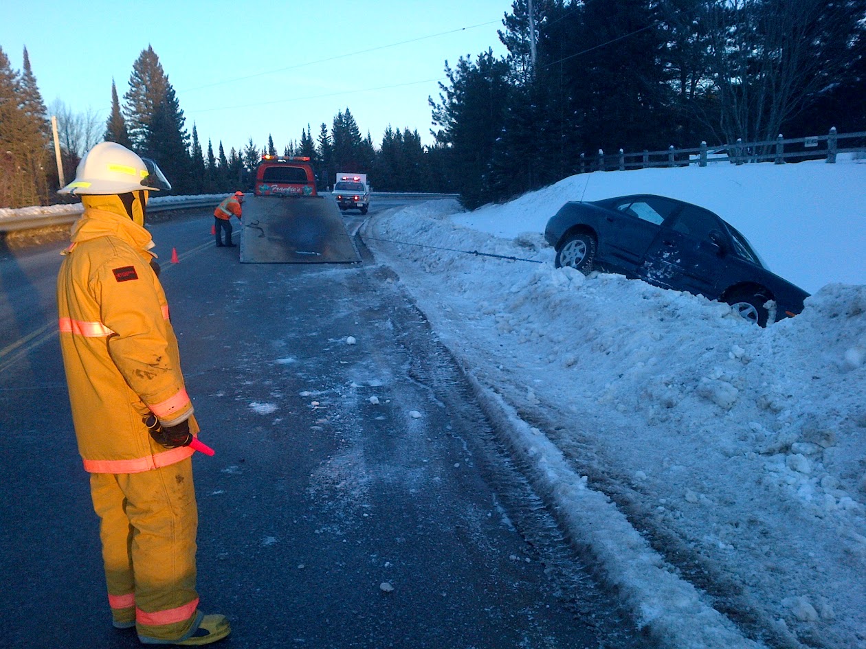 Firefighter directing traffic on a winter road while a tow truck and ambulance attend a car in the snowbank