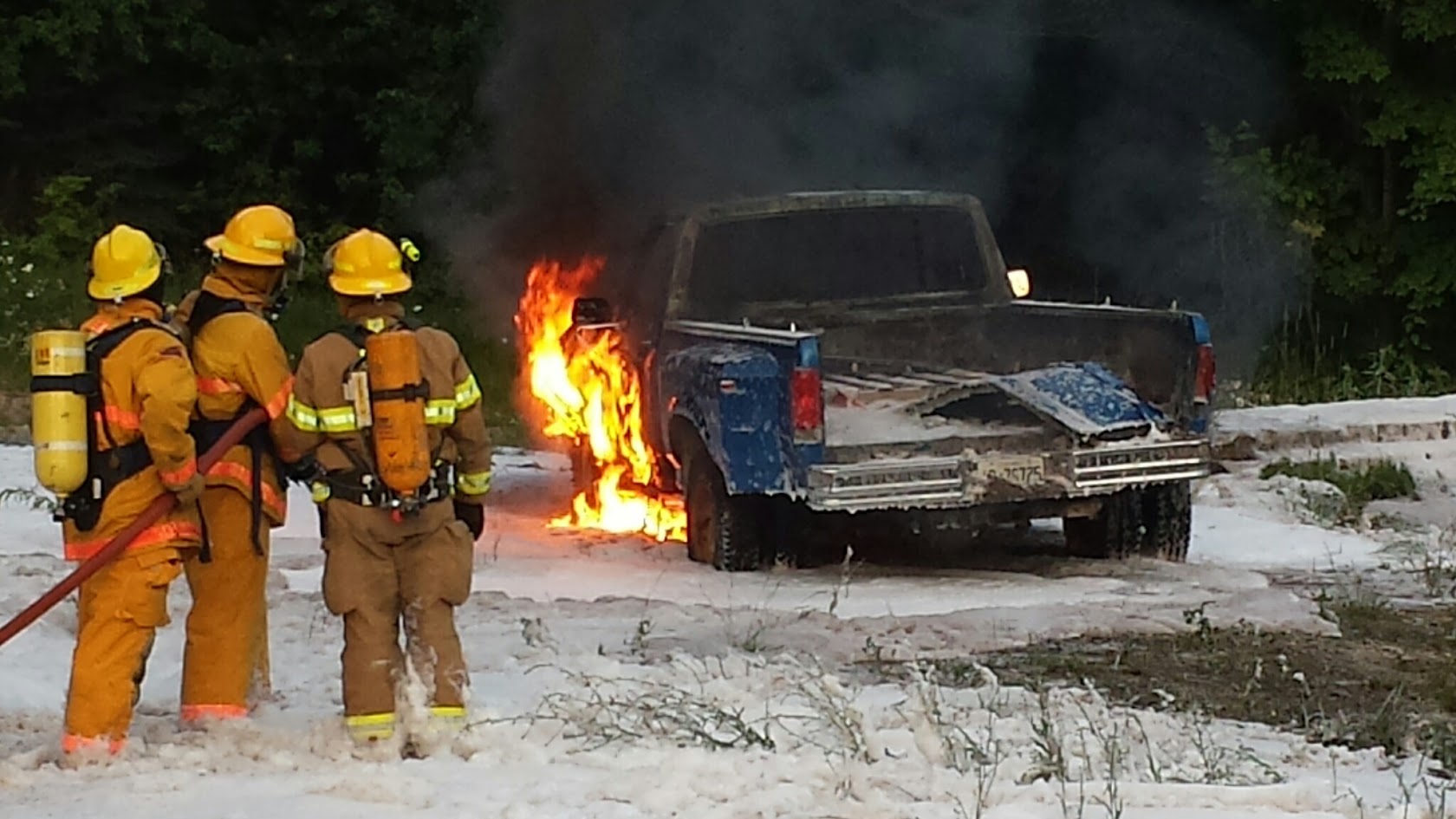 Three firefighters on air with a hose line at a burning pickup truck, ground covered in foam