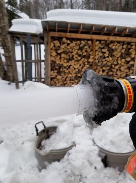 Gloved hand on a fire hose nozzle coated in ice, with water still flowing, beside a snow-covered woodshed