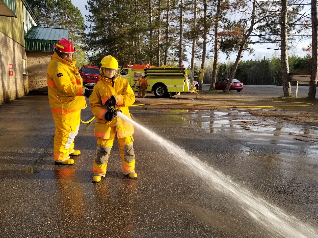 Young visitor in turnout gear operating a hose line with a firefighter beside them